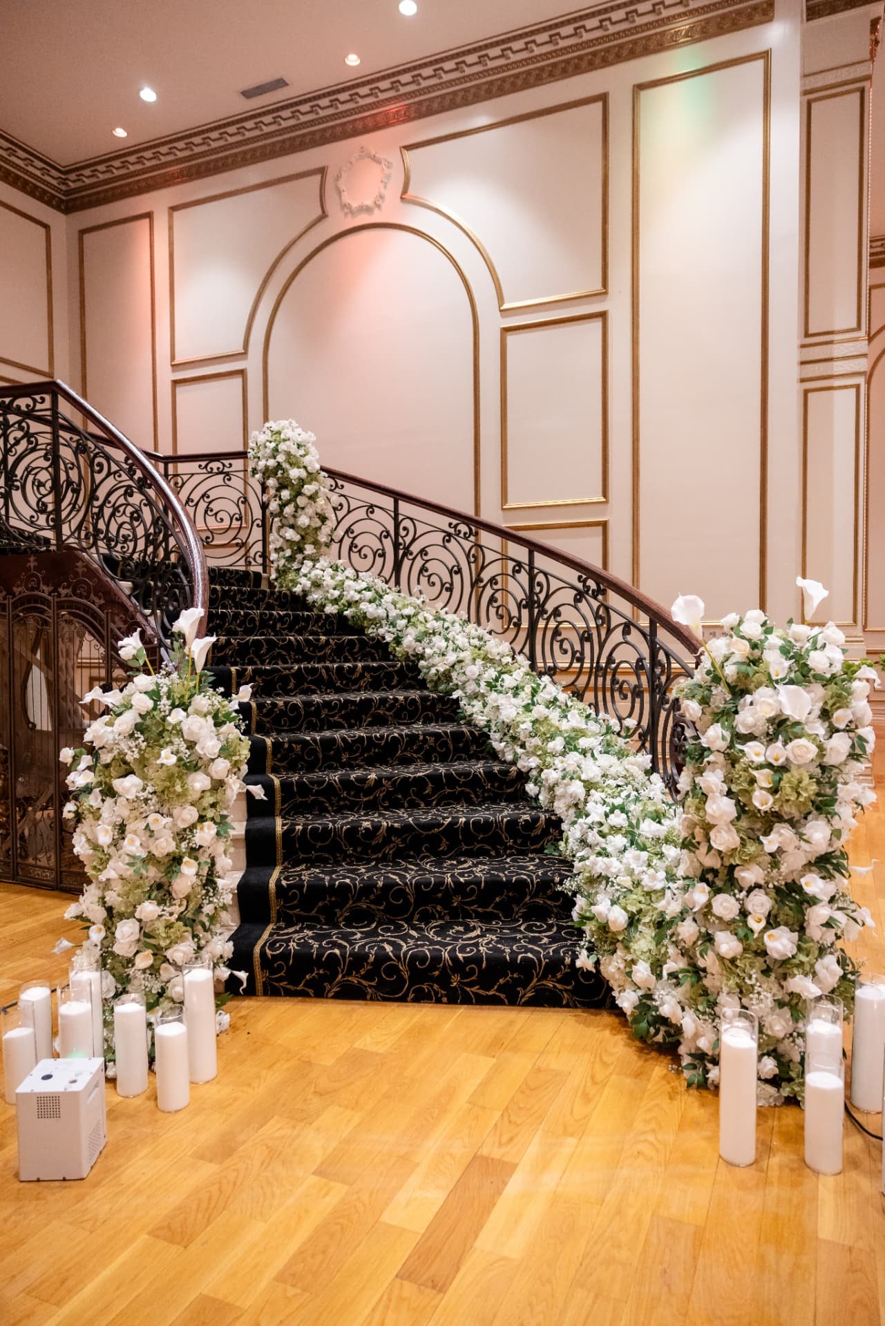 White rose and calla lily garland climbing a curved black and gold staircase lined with pillar candles at The Tides Estate in North Haledon, New Jersey, designed by Let's Bloom Florist