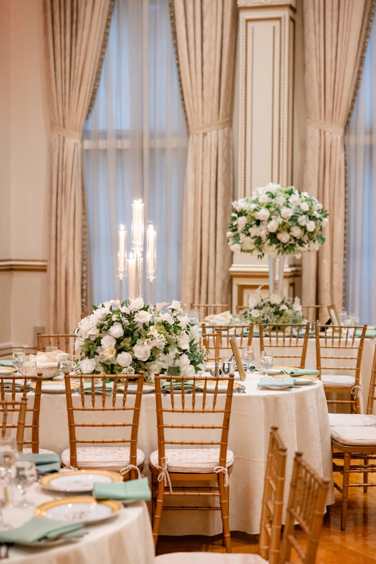 Round reception table with a white rose and calla lily arrangement, a clear candle tower and mint napkins before tall drapes at The Tides Estate in North Haledon, New Jersey, designed by Let's Bloom Florist